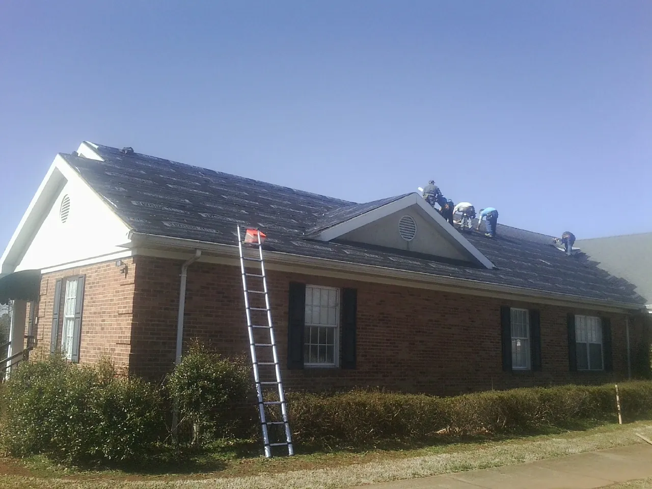 Crew tearing shingles off a brick ranch roof with a ladder set — Metal Roof Repair work by our crew in Elk Mound