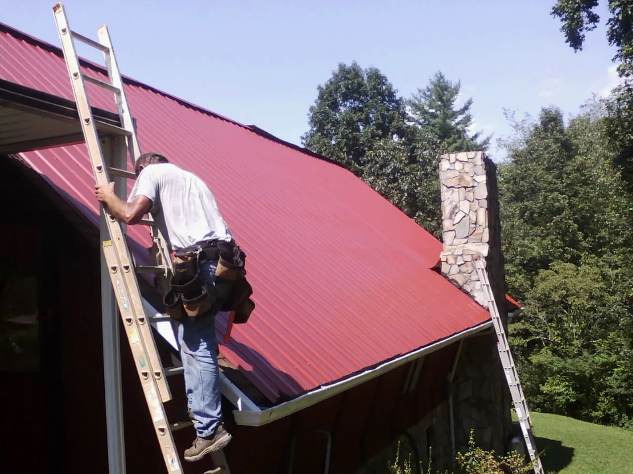 Roofer on a ladder beside a red metal roof and stone chimney — Soffit & Fascia Repair by Roofing Brothers in Elk Mound